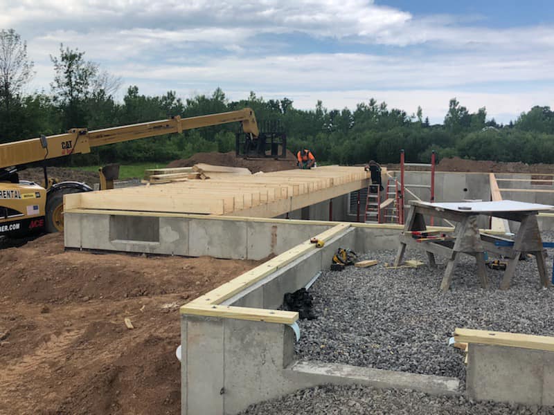 New home construction showing workers setting floor joists on a concrete foundation with equipment and materials on site.