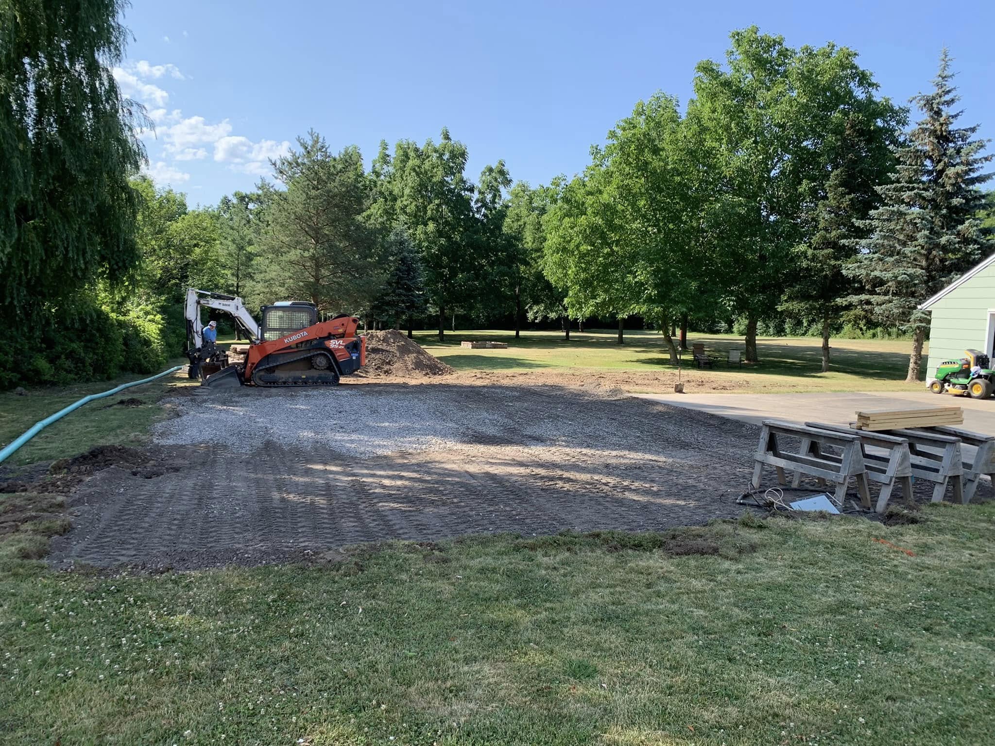 Site preparation with gravel base, skid‑steer loader, and excavator working on foundation area for new structure.