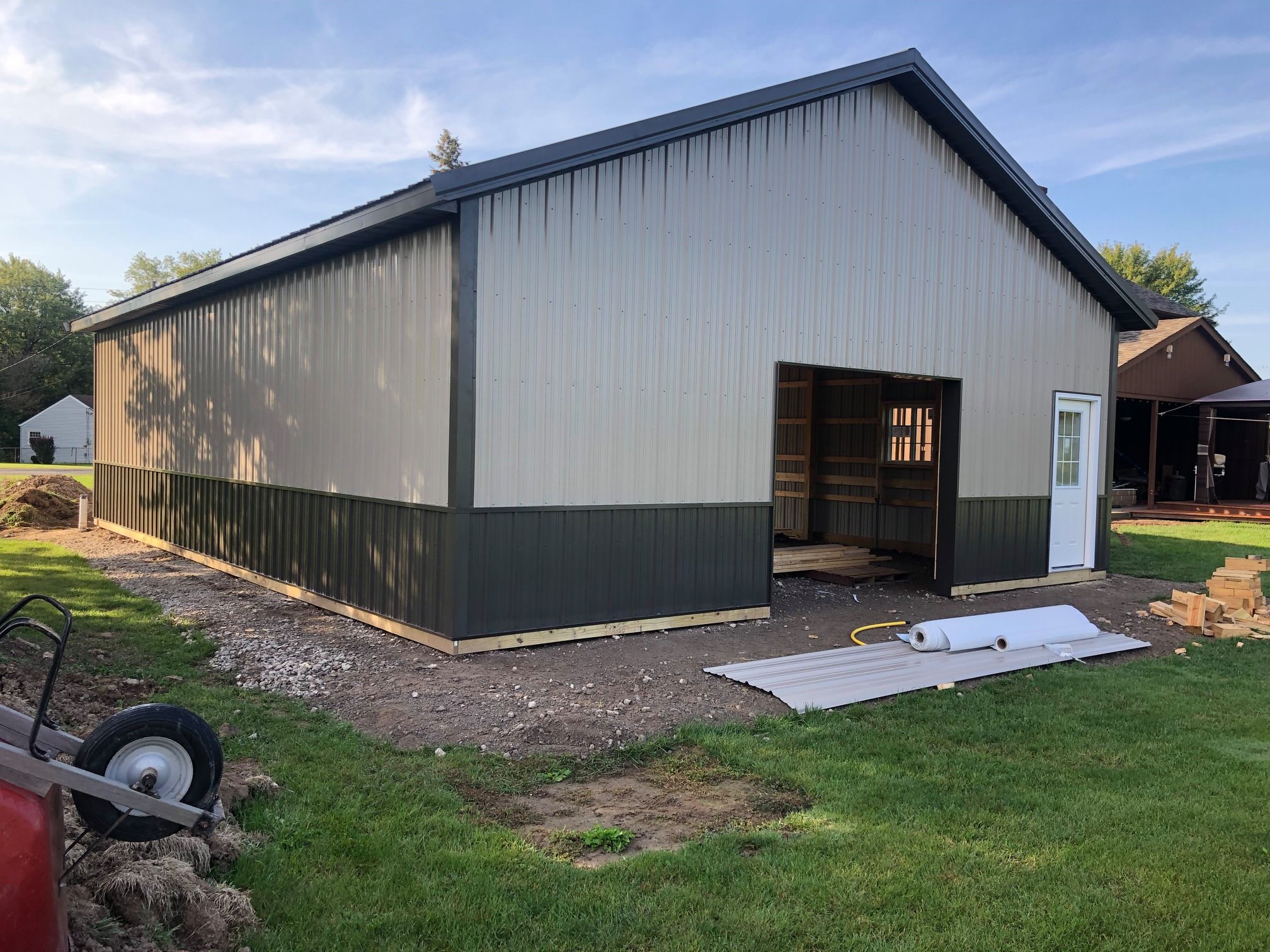 Newly constructed Post-Frame metal building with beige and green siding, open front entry, and construction materials on site during final stages of exterior installation.