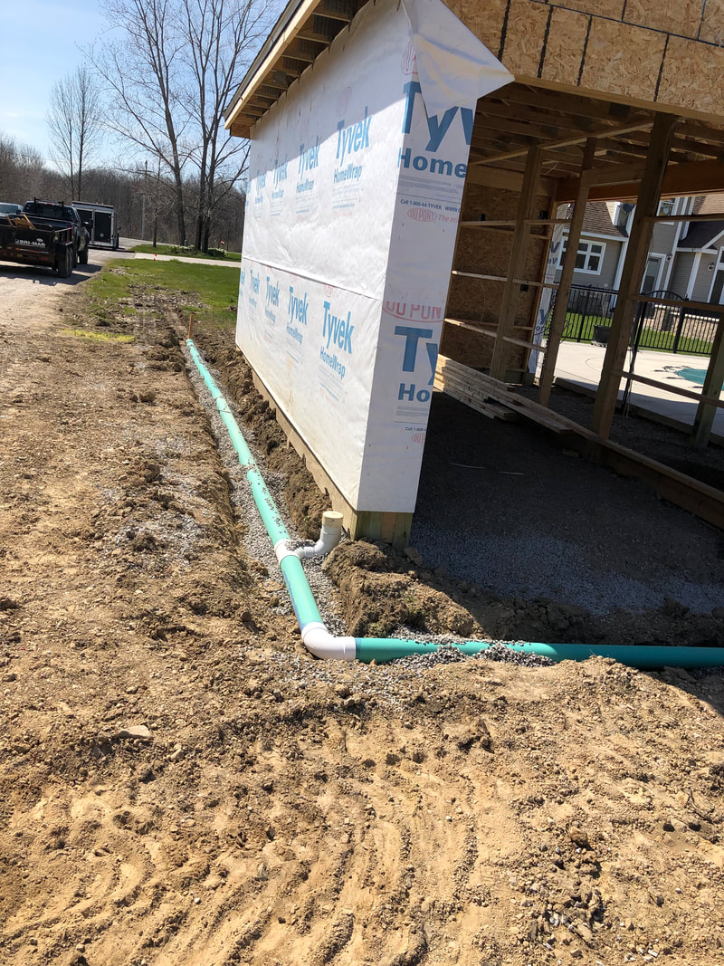 Green PVC drainage pipes laid in gravel trench beside Tyvek‑wrapped building during construction.
