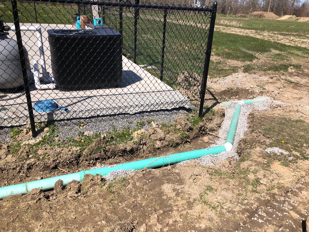 Drainage pipes installed near fenced pool equipment area, set in gravel trench during site excavation.